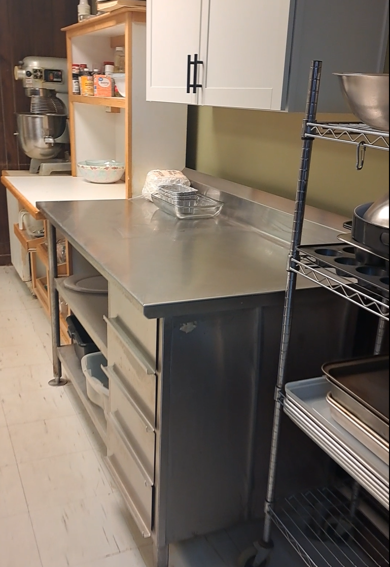 Kitchen prep area with stainless counter, mixer, shelving, and baking pans.
